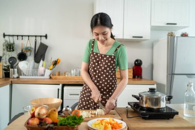 Asyalı genç bir kadın sabah evde mutfakta sağlıklı yemekler pişiriyor. Çekici kadın ev hanımı kendini mutlu hissediyor ve evde kahvaltıda çorba yapmak için malzemeler hazırlamaktan zevk alıyor..