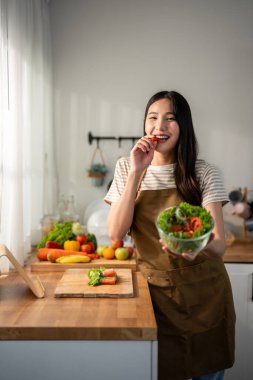 Asyalı kadın portresi evde mutfakta yeşil sağlıklı salata pişiriyor. Önlük giyen güzel bir genç kız, sabahları evdeki sağlık hizmetleri için temiz yemek yapmaktan zevk alıyor ve gülümsüyor, kameraya bakıyor..