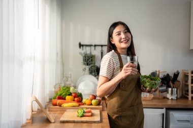 Asyalı kadın portresi sağlıklı salata yiyor ve temiz su içiyor. Önlük giyen güzel bir kız, sabahları evde sağlık hizmetleri için güzel yemekler yapmaktan zevk alıyor ve gülümsüyor, kameraya bakıyor..