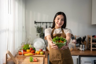 Asyalı kadın portresi evde mutfakta yeşil sağlıklı salata pişiriyor. Önlük giyen güzel bir genç kız, sabahları evdeki sağlık hizmetleri için temiz yemek yapmaktan zevk alıyor ve gülümsüyor, kameraya bakıyor..