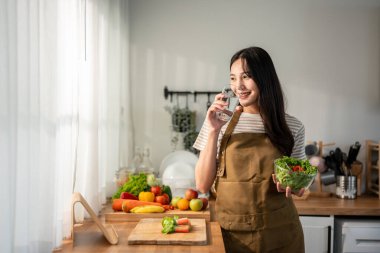 Asyalı kadın evde sağlıklı salata yiyor ve mutfakta su içiyor. Önlük giyen genç ve güzel bir kız, sabahları evdeki sağlık hizmetleri için temiz gıdalar yapmaktan zevk alıyor. Diyet ve Sağlıklı Yemek Konsepti.