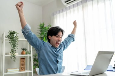 Asyalı heyecanlı serbest çalışan adam evde çalışan dizüstü bilgisayarını kullanıyor. Yakışıklı erkek öğrenci, evdeki oturma odasında başarılı bir şekilde iletişim kurmak için bilgisayar defterine yazıp sohbet ediyor..