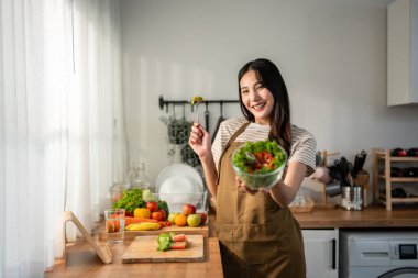 Asyalı kadın portresi evde mutfakta yeşil sağlıklı salata pişiriyor. Önlük giyen güzel bir genç kız, sabahları evdeki sağlık hizmetleri için temiz yemek yapmaktan zevk alıyor ve gülümsüyor, kameraya bakıyor..