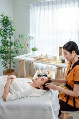 Asyalı genç bir kadın güzellik merkezinde kafa ve yüz masajı yaptırıyor. Çekici kız masaj yatağında uzanıyor, masaj kliniğinde vücut bakımı için masözden rahatlatıcı fizyoterapi hizmeti alıyor..