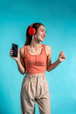 Kulaklık takan Asyalı bir kadın, telefonu mavi arka planda tutuyor. Çekici genç kız model mutlu ve neşeli hisset. Akıllı telefon cihazını kullan ve stüdyoda müzikle dans et..