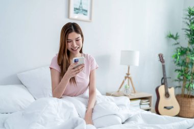 Asyalı çekici bir kadın sabah evde cep telefonu kullanıyor. Güzel bir kadın yatak odasında oturup hafta sonunun tadını çıkarıyor, akıllı telefonlardan evdeki internet teknolojisiyle sohbet ediyor..