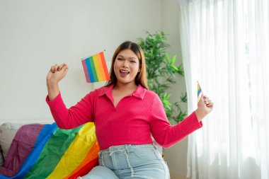 Elinde gökkuşağı bayrağı olan Asyalı trans kadın portresi, günlük hayatın tadını çıkarıyor. Çekici, kendine güvenen, gülümseyen ve kameraya bakan LGBTQ kadını tatillerini evde, oturma odasında geçirir..