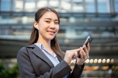 Şehirdeyken kulaklık takan Asyalı iş kadınının portresi. Çekici bayan çalışan mutlu ve rahat hissediyor, cep telefonuyla dışarıda yazıyor ve işten dinleniyor..