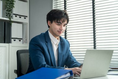 Ofiste çalışan Asyalı çekici iş adamı. Genç profesyonel erkek işçi mutlu ve rahat hissediyor. Şirket bölümündeki dizüstü bilgisayar planı projesini kullanarak masada oturuyor..