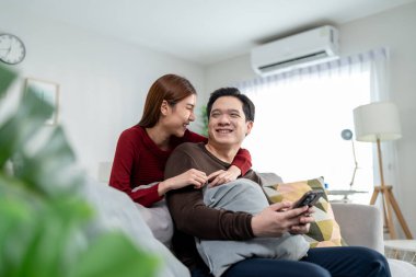 Asyalı genç çift evdeki rahat oturma odasında akıllı telefon kullanıyor. Çekici romantik yeni evli erkek ve kadın kanepede oturuyor, boş zamanlarını evde telefonda birlikte yazarak geçiriyorlar..