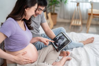 Asyalı genç çift bebeklerinin ultrasonunu dijital tabletten izliyor. Çekici, hamile bir koca ve hamile bir eş yatak odasında oturuyor, heyecanlanıyor ve doğmamış bir çocuğun görüntüsüne bakıyor..