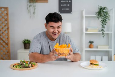 Asyalı büyük boy genç adam düşünceli bir şekilde taze salata ya da fast food 'u seçiyor. Çekici kıvrımlı erkek rahat bir oturma odasında oturmuş, dikkatli ve sağlıklı beslenme alışkanlığıyla hafta sonunun tadını çıkarıyor.