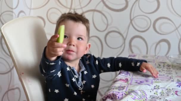 Child eating a peeled cucumber at a table in the kitchen — Wideo stockowe