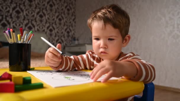 El niño dibuja felizmente mientras está sentado en la mesa de los niños — Vídeo de stock