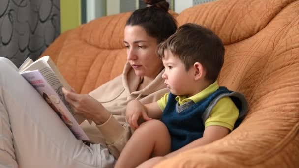 Matka z synem czytają książkę informacyjną siedząc na kanapie — Wideo stockowe