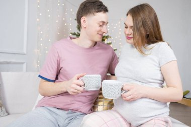 Arka planda Noel ağacı ışıkları varken evde çay ya da kahve içen mutlu çift. Tatiller, eğlence ve insanlar konsepti.