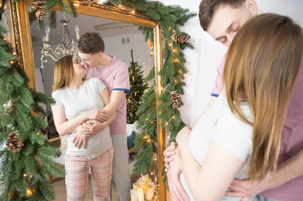 Koca, hamile karısına nazikçe sarılıp öpüyor ve arka planda Noel süsleri var..