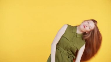 Uzun, güzel kızıl saçlı genç bir kadın kameraya bakarken başını sallıyor ve saçlarını sallıyor. Sarı arkaplan