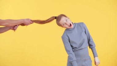 Erkek eller kızın at kuyruğunu çekiyor. Çilli genç bir kadın kameraya şaşkınlıkla bakıyor. Sarı arkaplan