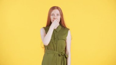 Çilli ve uzun kızıl saçlı genç bir kadın kameraya bakıyor. Ağzının fermuarını çekip çerçeveden uzaklaşıyor. Sarı arkaplan