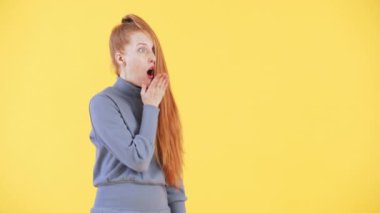 Yüzünde çilli ve kızıl at kuyruklu bir kadın kameraya bakıyor, şaşırmış bir yüz yapıyor ve bir şeyi işaret ediyor. Sarı arkaplan