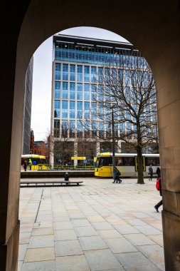 18 Şubat 2026 'da Manchester' da tasvir edilen Metrolink tramvay durağına ve modern ofis binalarına bakan taştan bir kemer deliğinden mimari bir manzara.