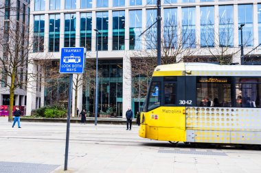18 Şubat 2026 'da şehir merkezindeki cam cephe ofis binasının önünden geçen sarı Manchester Metrolink tramvayı görüntüsü..