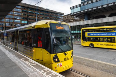 Parlak sarı bir Bee Network Metrolink tramvayı, 18 Şubat 2026 tarihinde arka planda sarı çift katlı bir otobüsle Manchester 'daki bir istasyon peronuna yanaşıyor..