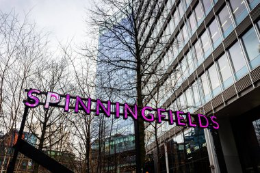 Mor neon renkli Spinningfield ilçe tabelası, arka planda 18 Şubat 2026 tarihinde resmedilen modern cam ofis binaları olan ağaçlar arasında yer almaktadır..