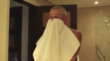 Bir adam aynanın önünde beyaz bir havluyla yüzünü siliyor.