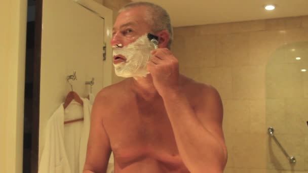 Een man scheert zijn baard met een scheermes terwijl hij voor een spiegel staat — Stockvideo
