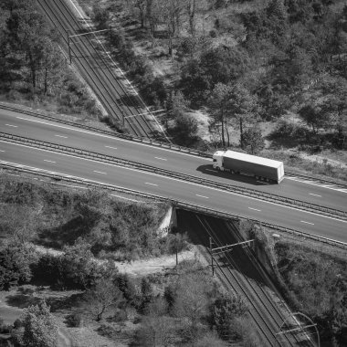 Kamyonlar otoyol ve demiryolunu birbirine bağlayan bir üst geçitten geçiyor. Etrafı ağaçlarla çevrili. Sahnenin alt kısımlarında birkaç ev görünüyor..