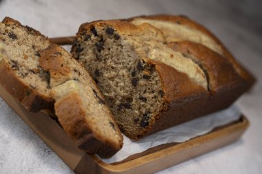 Freshly baked banana bread is sliced and displayed on a wooden cutting board. The bread has a golden-brown crust and chocolate chips, perfect for a cozy snack at home.