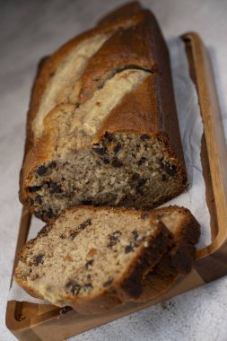 Freshly baked banana bread is sliced and displayed on a wooden cutting board. The bread has a golden-brown crust and chocolate chips, perfect for a cozy snack at home.