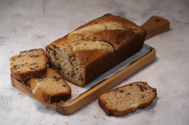 Freshly baked banana bread is sliced and displayed on a wooden cutting board. The bread has a golden-brown crust and chocolate chips, perfect for a cozy snack at home.