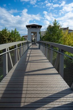Mont de Marsan 'daki yeşillik boyunca uzanan ahşap bir köprü ziyaretçileri beyaz bir kemer yoluna yönlendiriyor. Parlak bulutlar gökyüzünü dolduruyor, bu kentsel alanın sakin atmosferini güçlendiriyor..