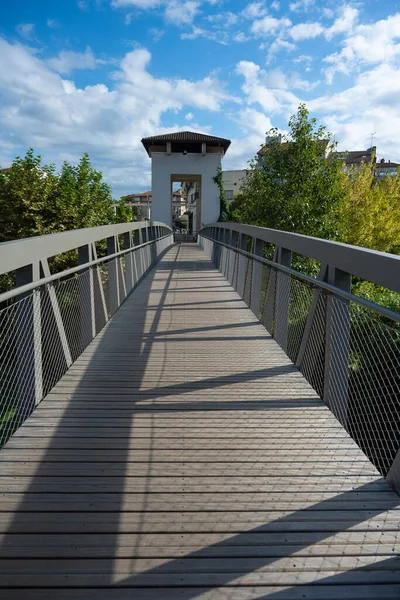 Mont de Marsan 'daki yeşillik boyunca uzanan ahşap bir köprü ziyaretçileri beyaz bir kemer yoluna yönlendiriyor. Parlak bulutlar gökyüzünü dolduruyor, bu kentsel alanın sakin atmosferini güçlendiriyor..