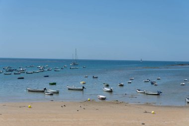 İspanya 'nın Endülüs kentinde, Cadiz sahilinin yaz manzarası, renkli şemsiyeler ve sakin deniz sularına demirlemiş teknelerle. Tatil, rahatlama, deniz, güneş ve Akdeniz atmosferi kavramları için mükemmel olan tipik İspanyol deniz kıyısı turizmi sahnesi.