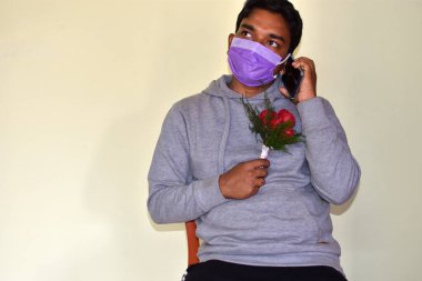 Coronavirüs salgını sırasında Sevgililer Günü. Çekici bir adam ameliyat maskesi takıyor cep telefonlu ve beyaz arka planda güller ve fotokopi alanı var. - Evet. Covid-19 yaşam tarzı