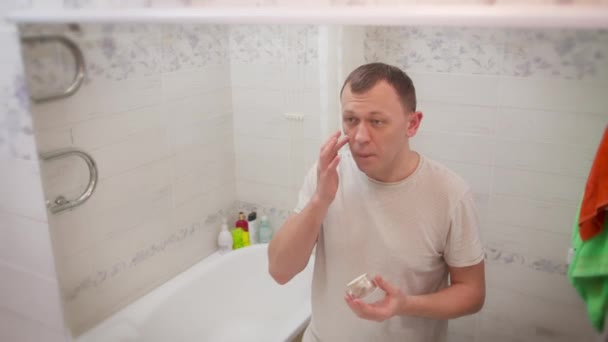 Genç adam yüz yüze makyaj kremi sürüyor, banyoda duruyor, cilt bakımı yapıyor. — Stok video