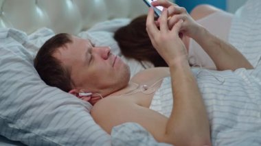 Aile yatakta dinleniyor, kadın gözlerini çevirmiş uyuyor, koca kulaklıkla müzik dinliyor.