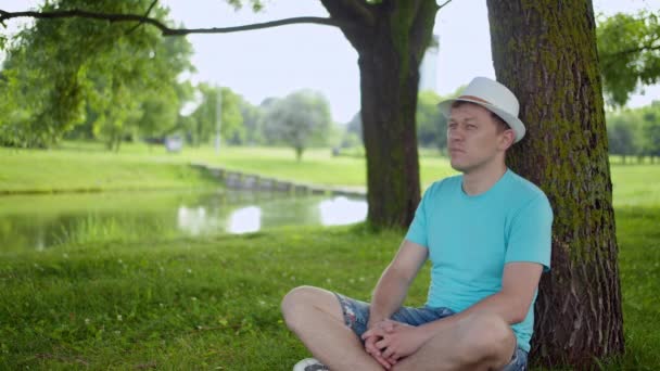 Homme attrayant en chapeau blanc reposant sur l'herbe sous l'arbre — Video