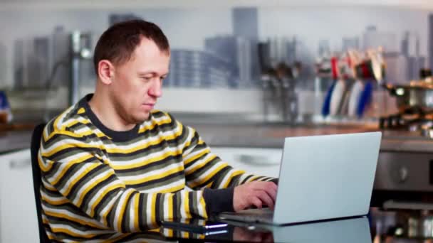 Male freelancer working on laptop at home while sitting at table, kitchen background — Stock Video