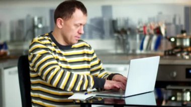 Evde dizüstü bilgisayarda çalışan serbest çalışan bir adam masada oturuyor, arka plan mutfağı...