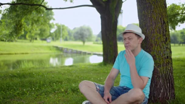 O jovem em um chapéu senta-se pensativamente na grama abaixo de uma árvore — Vídeo de Stock
