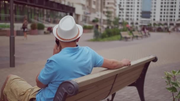 Un touriste homme en chapeau blanc se repose assis sur un banc de la ville, mouvement de la caméra, espace de copie — Video