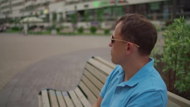 Giovane uomo in occhiali da sole sta riposando mentre seduto su una panchina della città, movimento della fotocamera, spazio copia — Video Stock