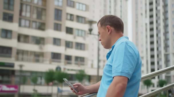 Fiatal férfi rövidnadrágban, mobiltelefonnal áll a lépcsőn, közel a korláthoz, a város hátterében. — Stock videók