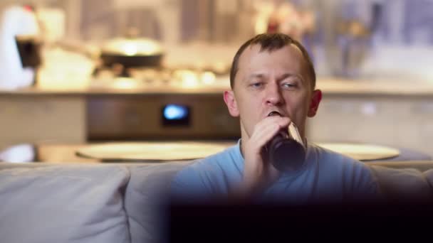 Mężczyzna pijący piwo z butelki, siedzący w domu na kanapie i oglądający telewizję, ruch kamery — Wideo stockowe