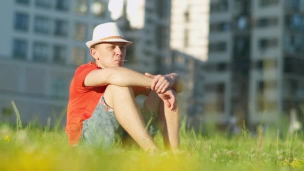 Man in hat is resting while sitting on the grass in a city park, tracking the camera — Stock Video